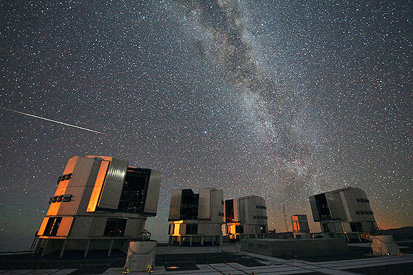Cielo nocturno con la Vía Láctea y una estela de meteoro sobre cúpulas de observatorio, tonos azul oscuro y estrellados